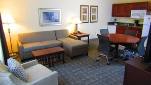 A living room with a sofa, a small table, a chair, a dining table with chairs, a kitchen area with cabinets and appliances, and framed pictures on the wall.