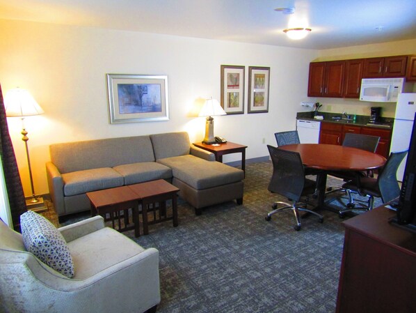 A living room with a sofa, a small table, a chair, a dining table with chairs, a kitchen area with cabinets and appliances, and framed pictures on the wall. - Queen Suite with In-Suite Conference Space Near the University of Rochester (Rochester)