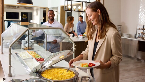 A buffet area with various food options, a glass display case, and a person serving food. - Queen Studio Suite | Full Kitchen Included (Lafayette)