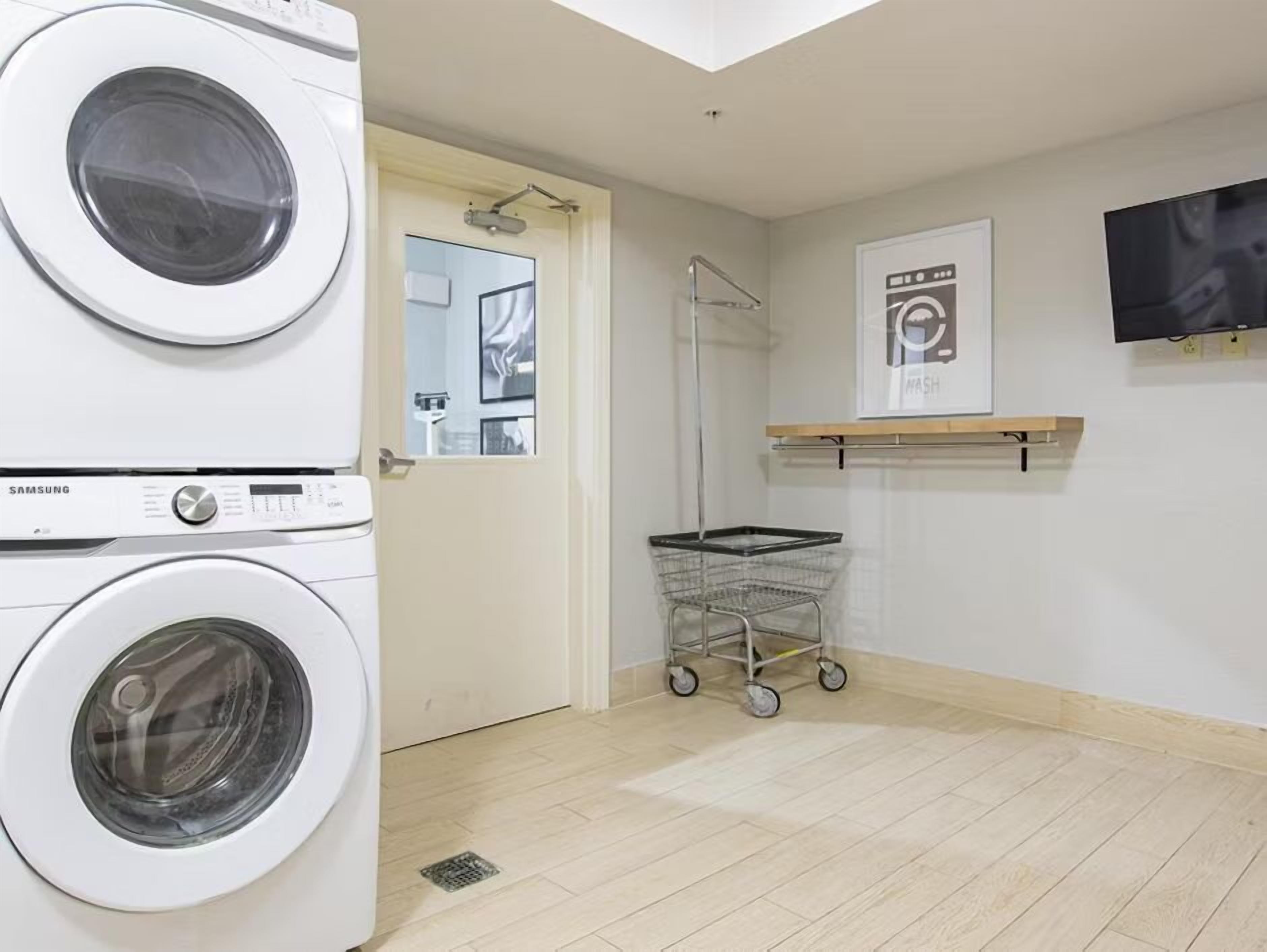 A laundry room with two white washing machines, a metal laundry cart, a door leading to a bathroom, and a wall-mounted TV.