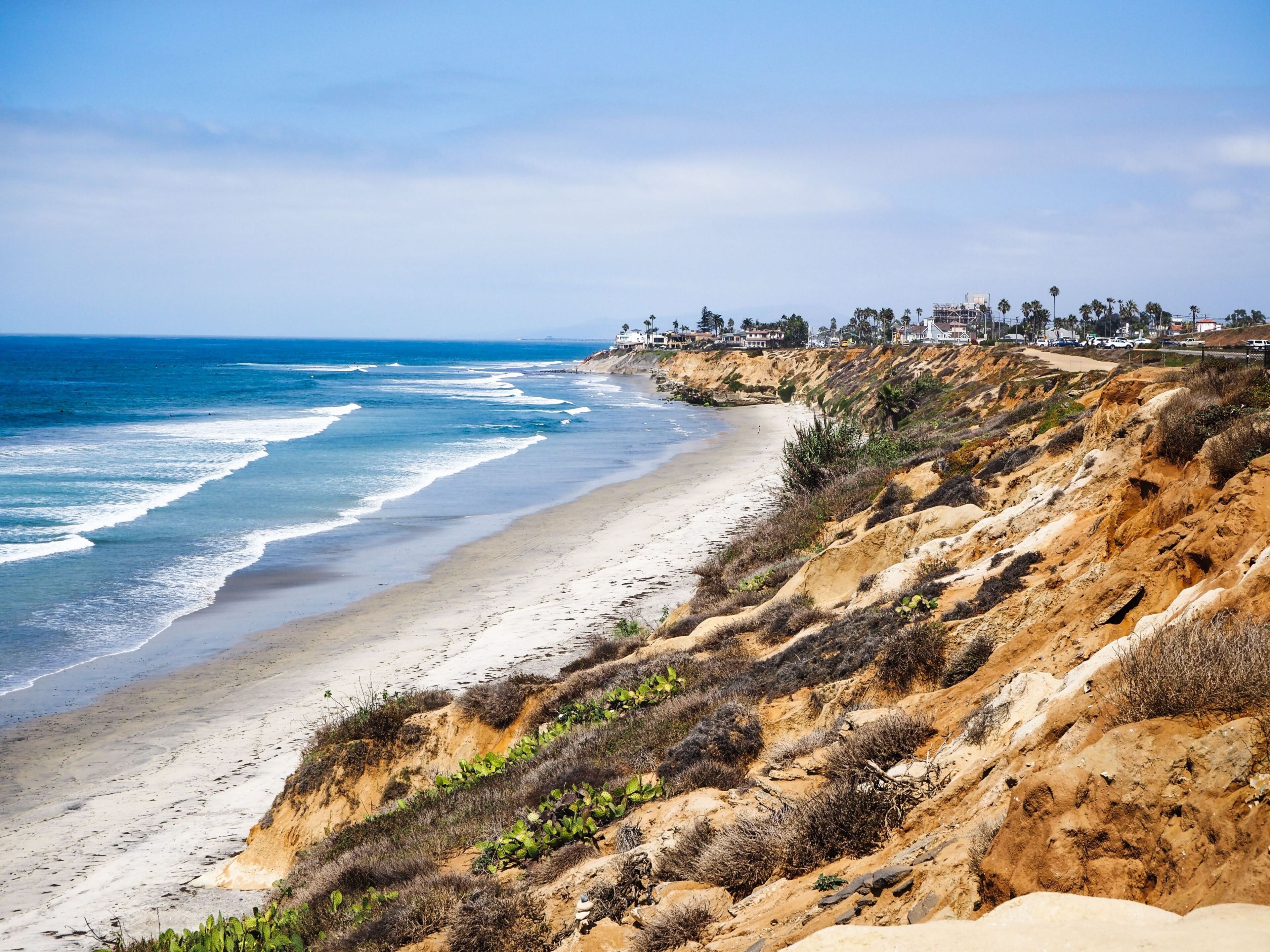 A coastal landscape with a sandy beach, rocky cliffs, and a distant residential area.