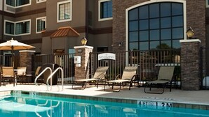 Une piscine entourée de chaises longues et de parasols, elle-même bordée d’un immeuble à plusieurs étages, doté d’une façade en briques et de grandes fenêtres.