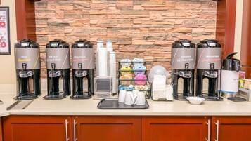 A breakfast bar with multiple coffee makers, a tray with cups, and a glass display of pastries and condiments.
