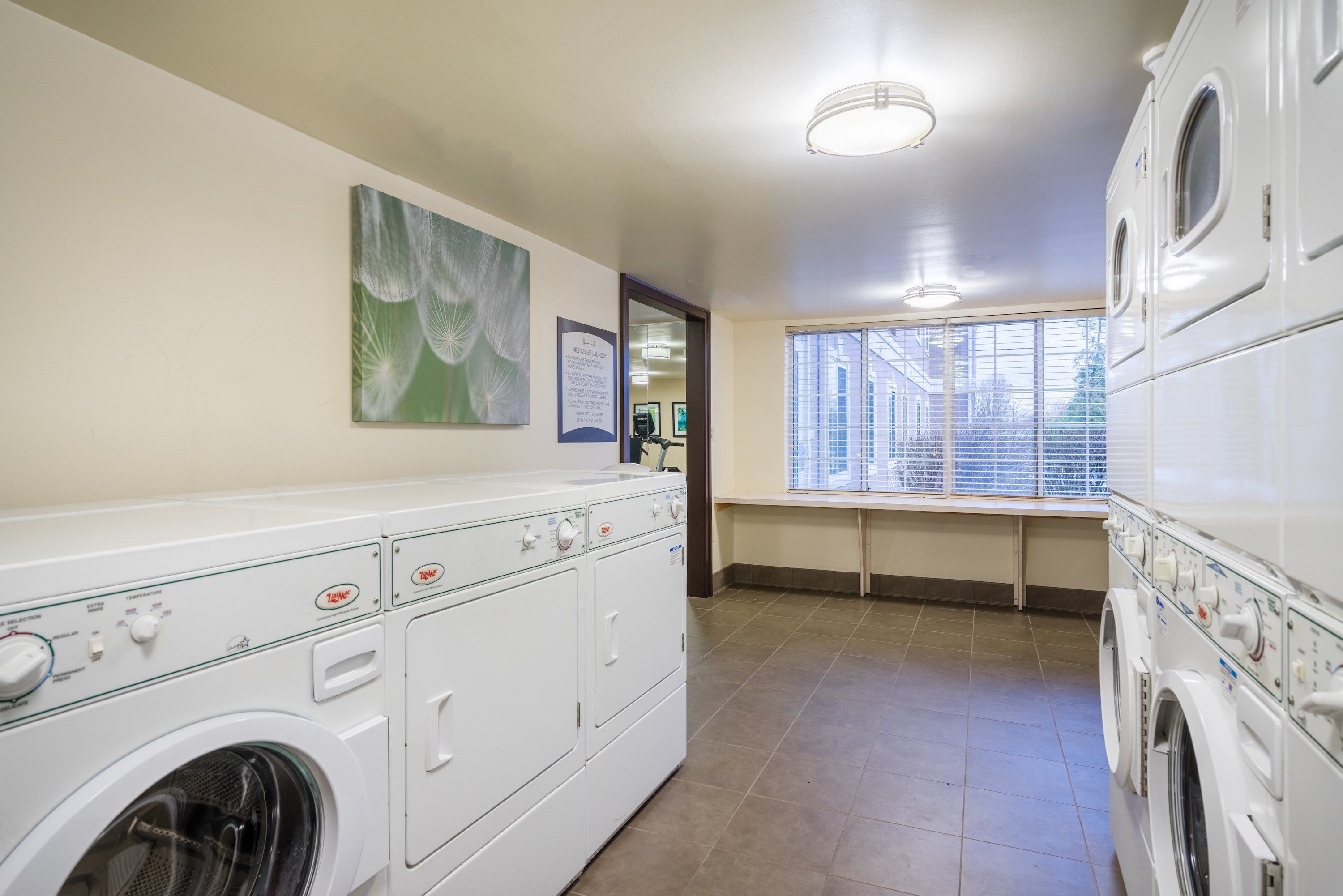 A laundry room with stacked washers and dryers, a window with blinds, and a ceiling light.