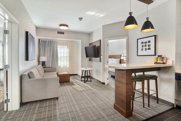 A modern hotel room with a sofa, a small table, a TV, and a kitchenette.