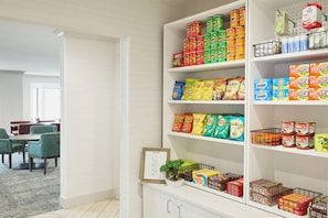 Eine Speisekammer mit Regalen voller verschiedener Lebensmittel, ein Essbereich mit grünen Stühlen und ein gerahmtes Bild auf einem Schrank.
