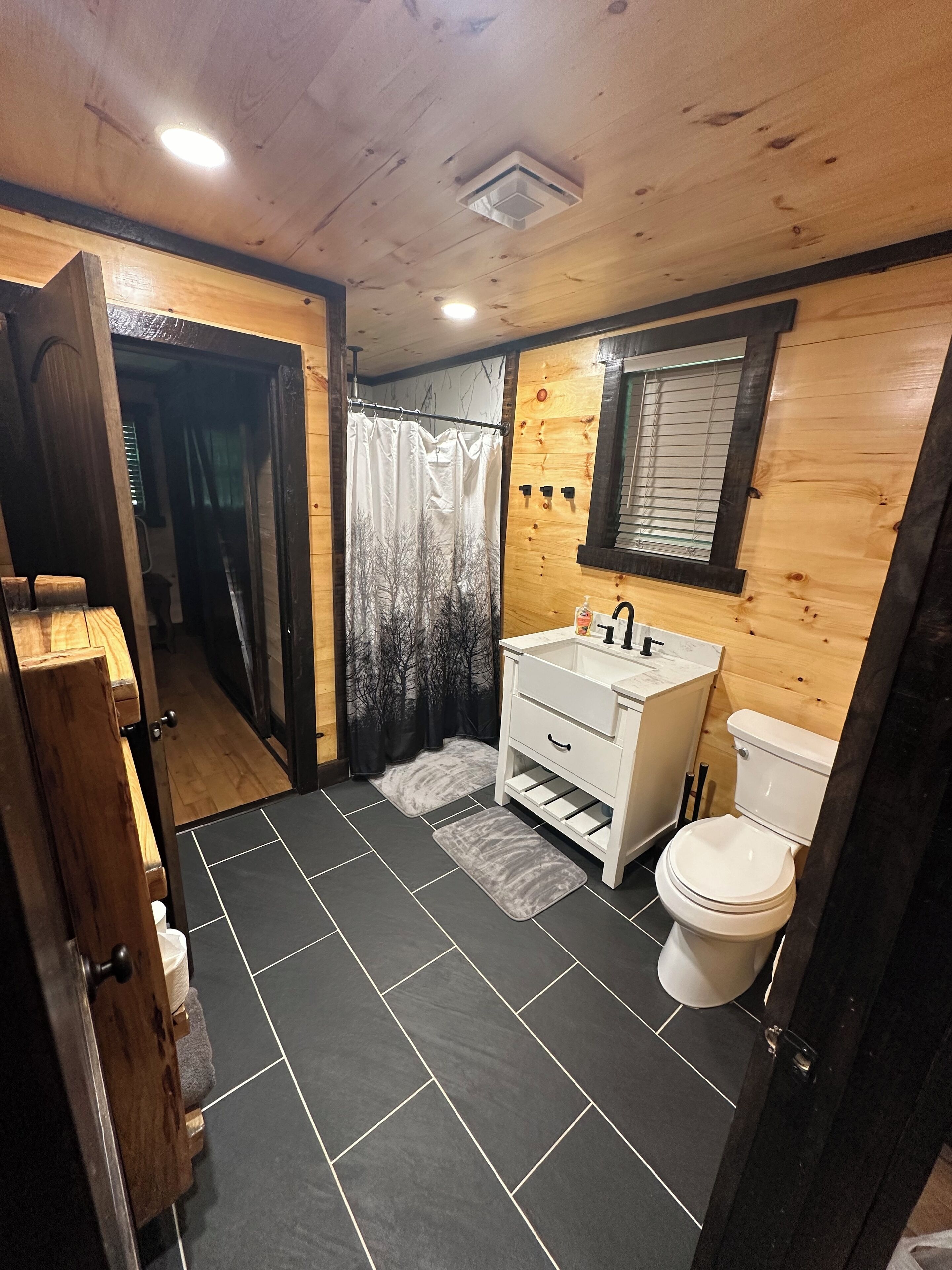 Un bany amb interior de fusta, un lavabo blanc, un vàter i una cortina de bany amb un disseny d'arbre.
