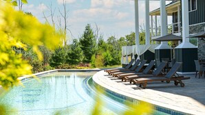 Un espace piscine agrémenté de chaises longues et de parasols, entouré de verdure et bordé d’un bâtiment aux éléments en pierre et en bois.