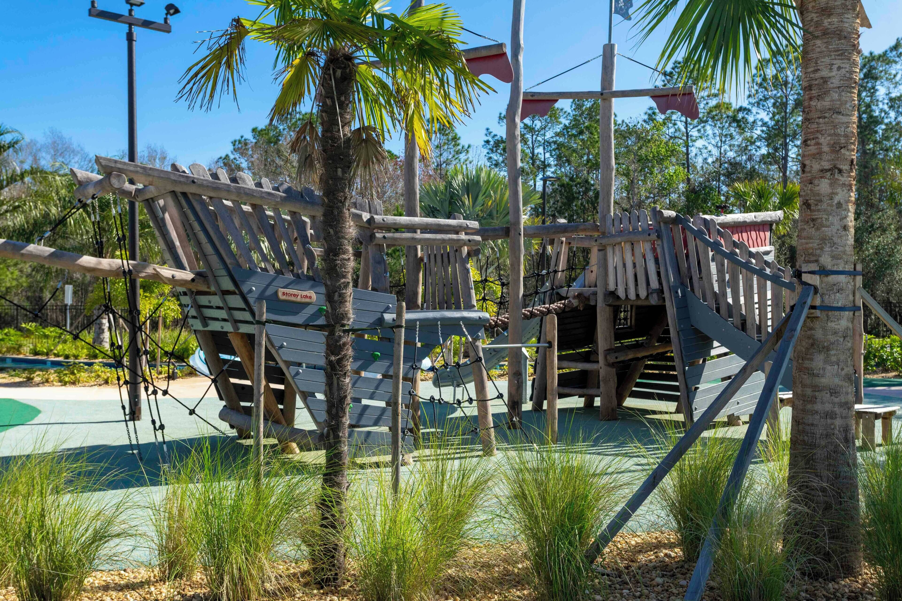 A wooden play structure with rope bridges and a slide, surrounded by palm trees and greenery.
