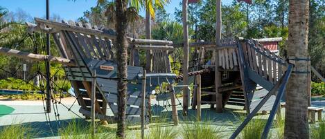 A wooden play structure with rope bridges and a slide, surrounded by palm trees and greenery.