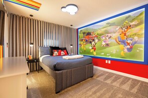 Un dormitorio con un mural grande de personajes animados, una cama con ropa de cama gris, una mesita de noche y un escritorio.