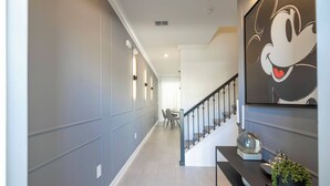 A hallway with a staircase, a dining area in the background, and a large Mickey Mouse painting on the wall. - Private 4BR Check In and Chill Out (2417ST) (Kissimmee)