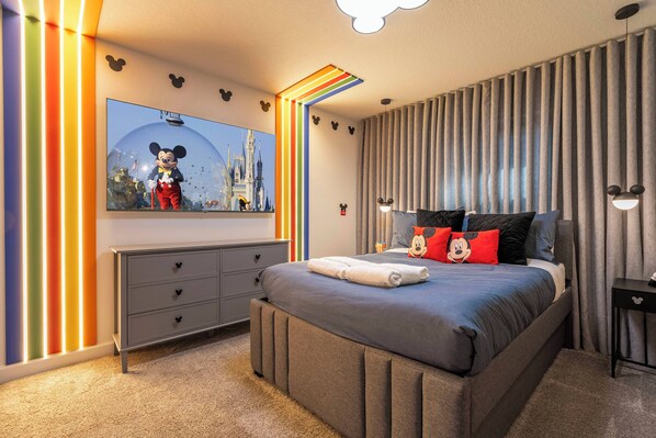 Un dormitorio moderno con una cama grande, un aparador, un muro con temática de Mickey Mouse y un amplio ventanal con cortinas.