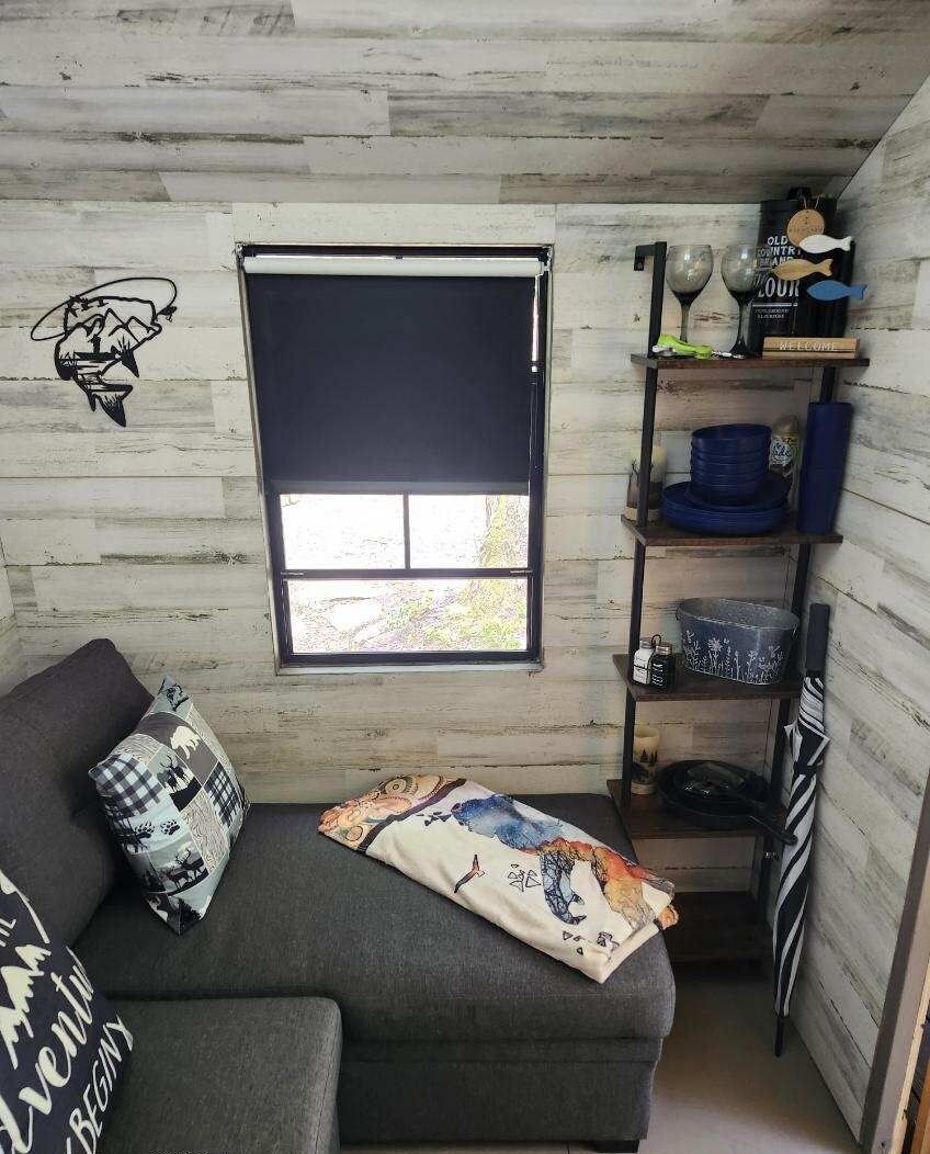 A cozy room with a grey sofa, a window with a black blind, and a shelf with various items including a blue bowl and a candle.