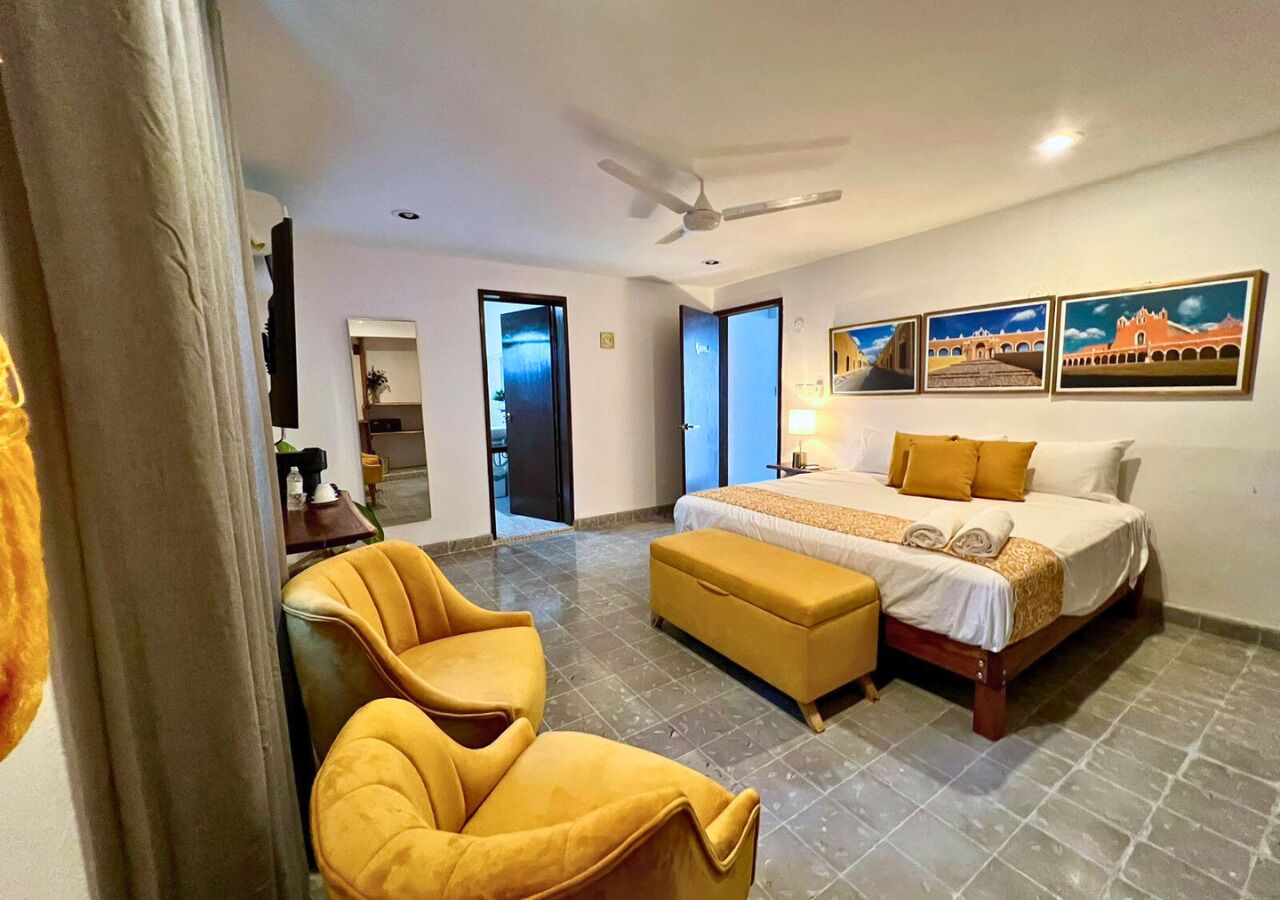 Habitación de hotel con cama, escritorio, silla y banqueta. Hay cuadros en la pared y un ventilador de techo.