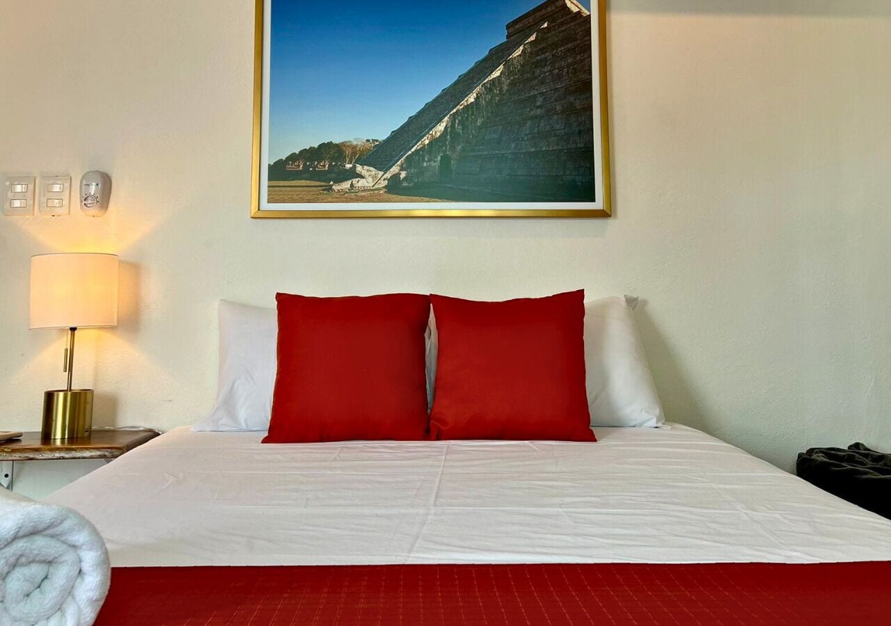 Una habitación de hotel con una cama grande, ropa de cama roja y blanca, una mesita de noche con una lámpara y un cuadro en la pared.