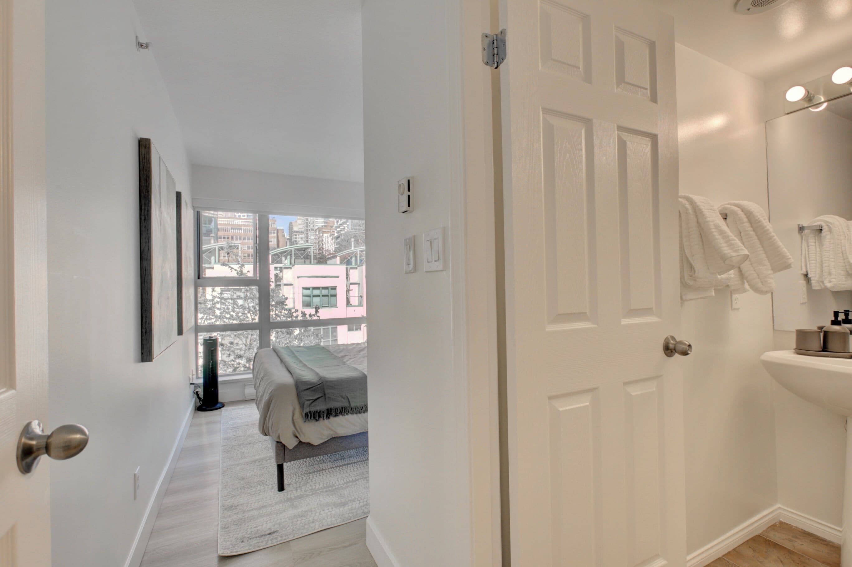 A hotel room with a large window, a bed, a white sink, and a door.