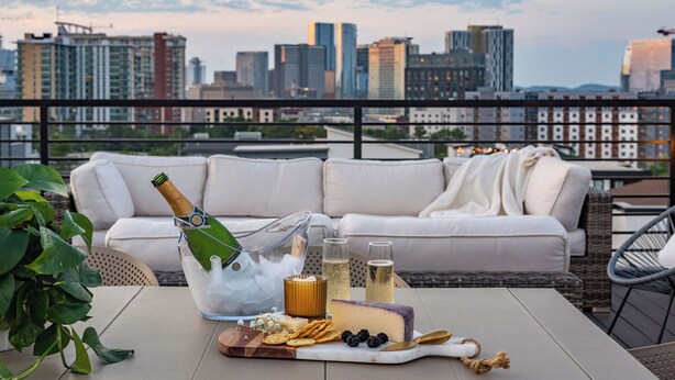 Una terraza en la azotea con vistas a un horizonte urbano, un sofá blanco, una mesa con una botella de champán, copas, queso, galletas y una planta.