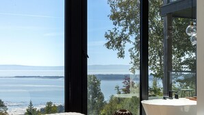A bedroom with a large window overlooking a body of water and mountains, a white bathtub, and a woven basket. - Le Lord, River view, trail and snowmobile (Les Éboulements)