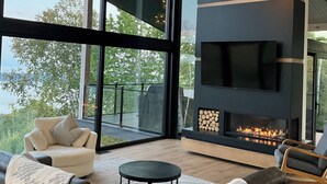 A modern living room with a fireplace, large windows, and a view of the outdoors. - Le Lord, River view, trail and snowmobile (Les Éboulements)
