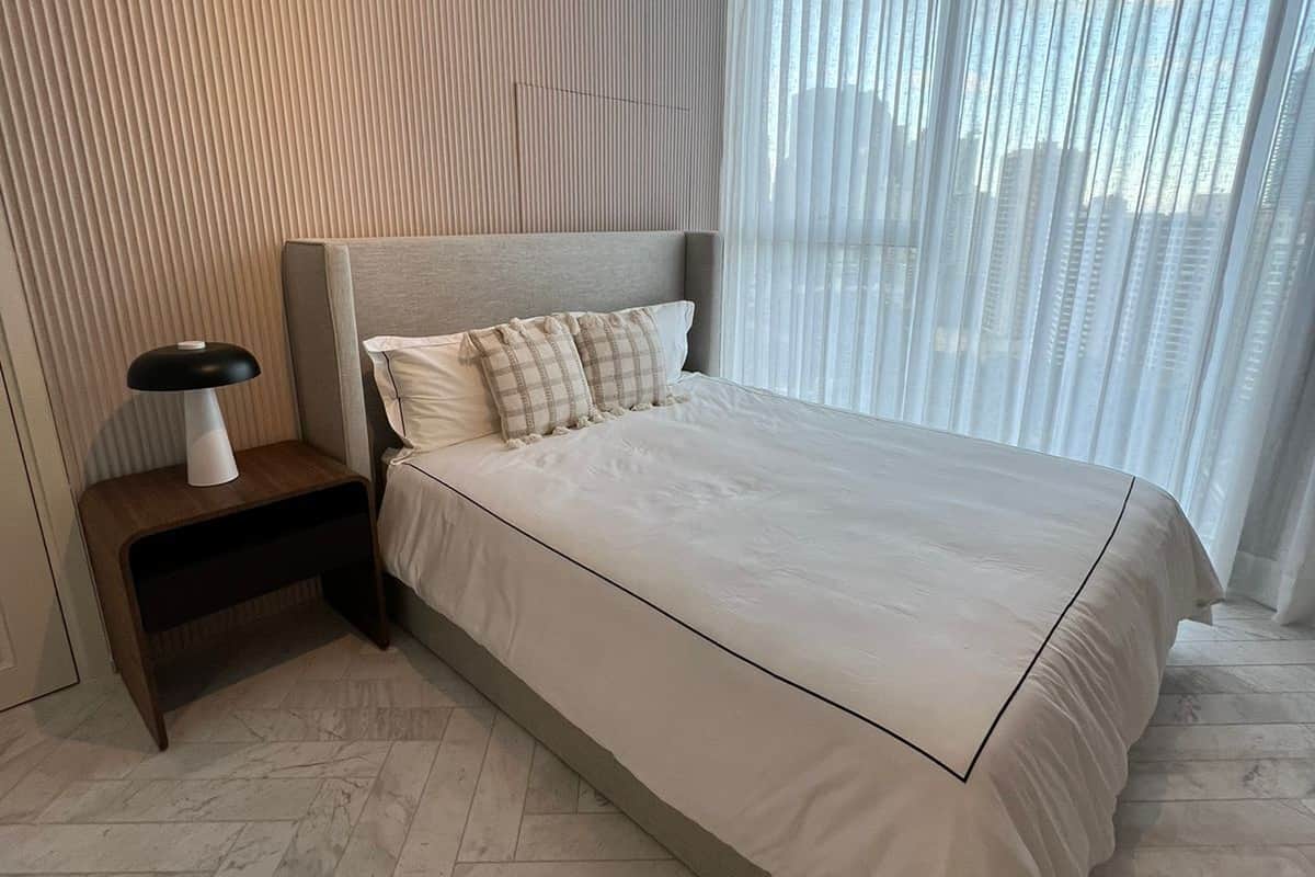 A modern bedroom with a large bed, a nightstand, and a window with sheer curtains.