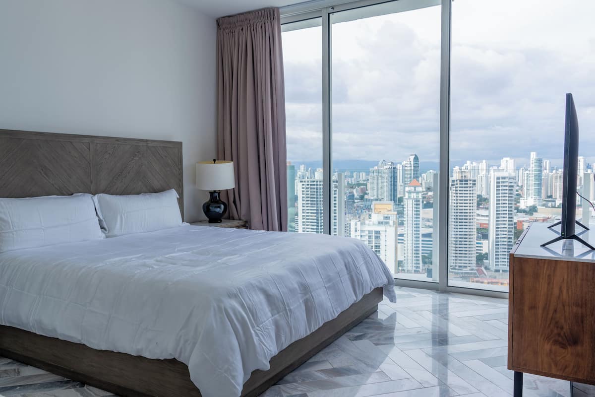 A hotel room with a large bed, a nightstand, a lamp, and a view of a cityscape through large windows.