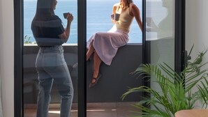 Ein Zimmer mit einem großen Bogenfenster mit Blick auf das Meer, einer Kühlpflanze und einem Holznachttisch.