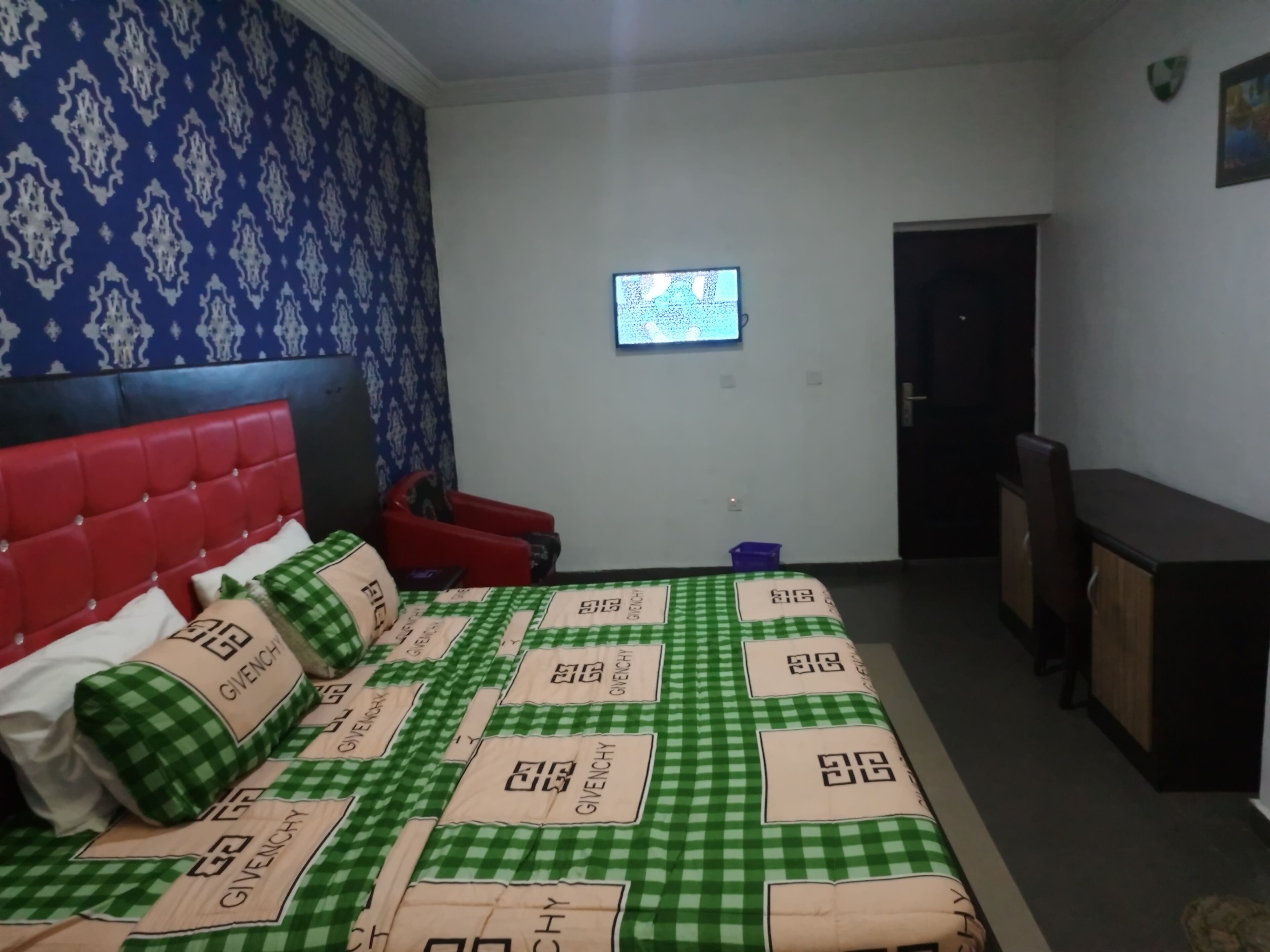 A bedroom with a red headboard, a bed with green and beige bedding, a small TV on the wall, and a door leading to another room.