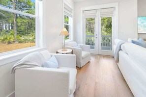 Une pièce avec un canapé blanc, une petite table ronde et une lampe. Une grande fenêtre offre une vue sur les arbres.