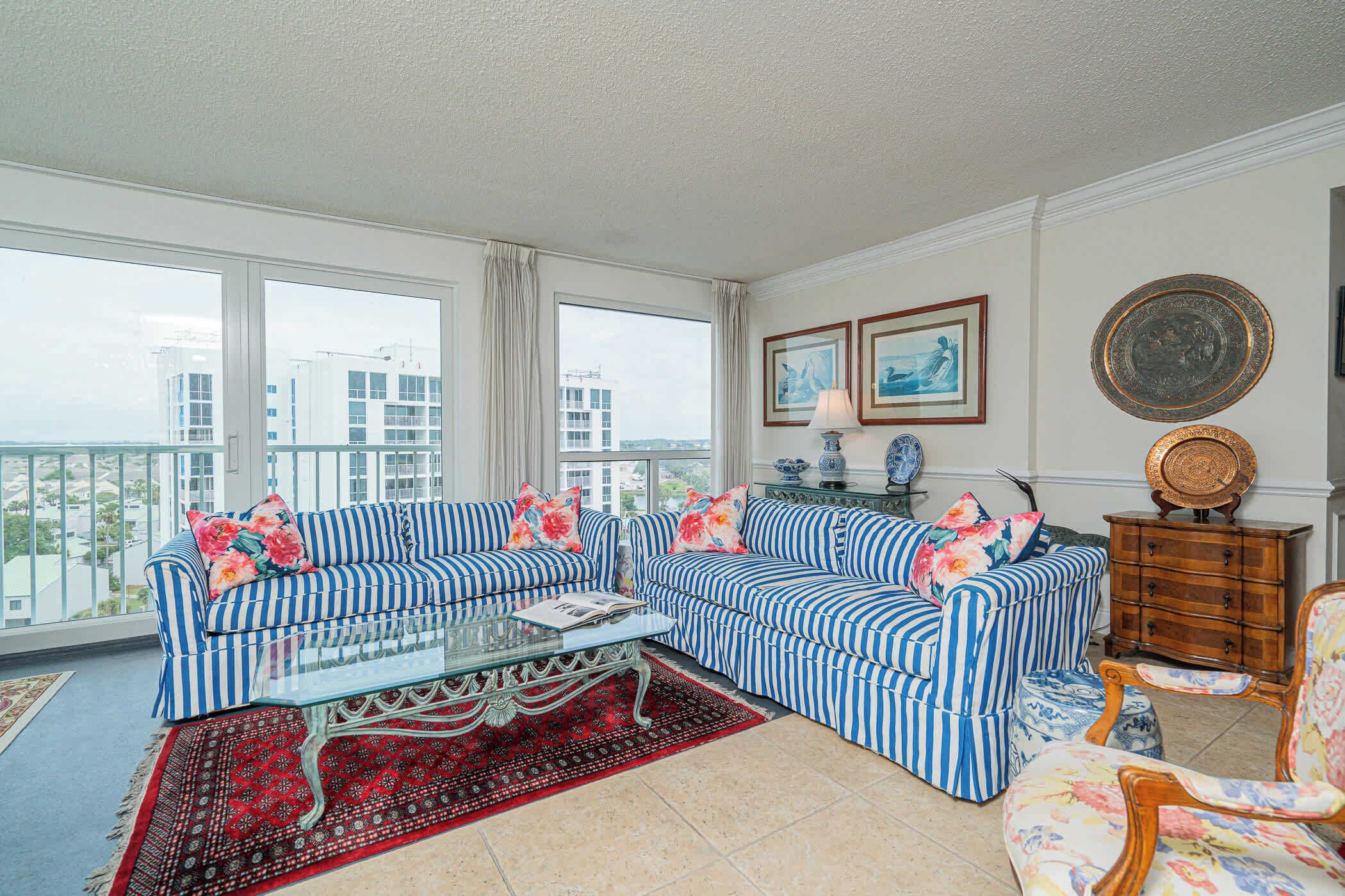 A living room with striped sofas, a glass coffee table, and a red patterned rug. There are two framed pictures on the wall and a wooden cabinet with decorative items.
