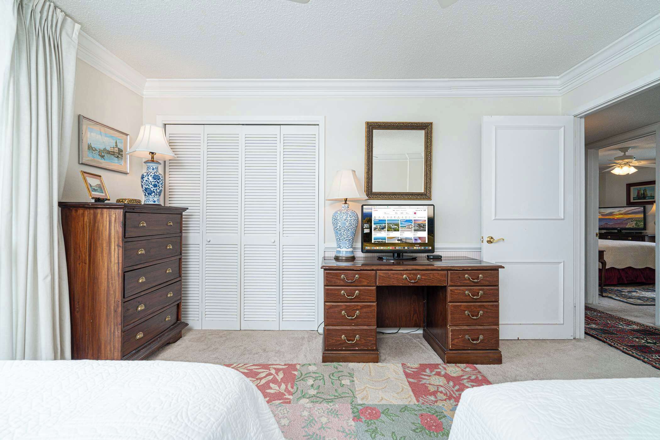 Une chambre avec deux lits, une commode en bois, un bureau avec un ordinateur, une télévision et une porte donnant sur une autre pièce.