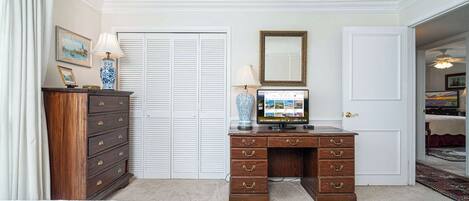 Une chambre avec deux lits, une commode en bois, un bureau avec un ordinateur, une télévision et une porte donnant sur une autre pièce.
