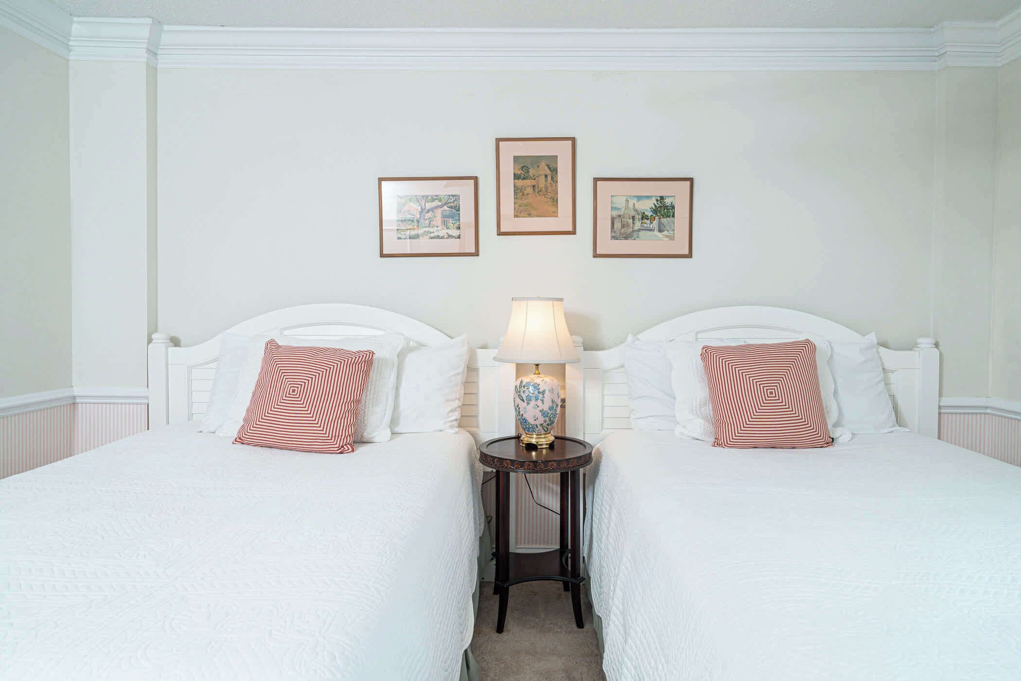 A hotel room with two beds, a small table with a lamp, and three framed pictures on the wall.