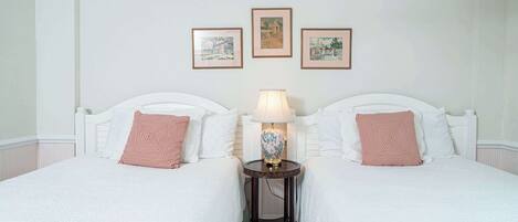 A hotel room with two beds, a small table with a lamp, and three framed pictures on the wall.