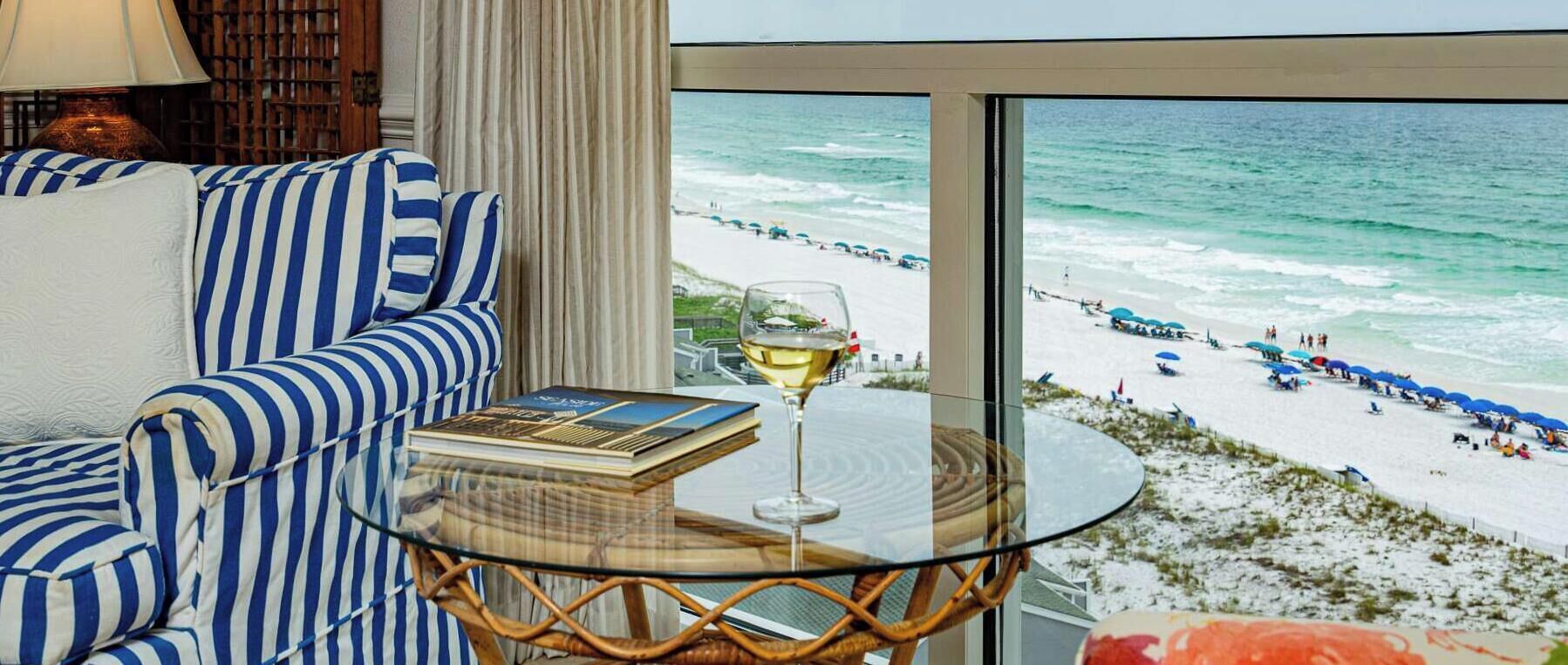 A room with a view of a beach, a glass table with a book and a glass of wine, and striped blue and white furniture.