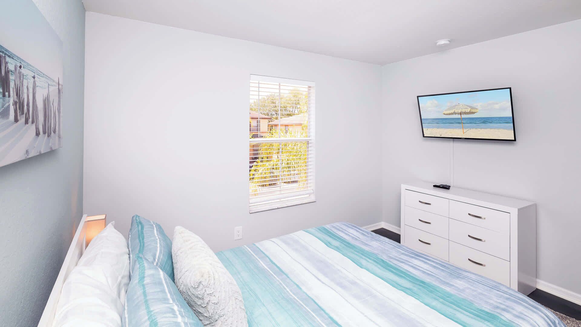 Een slaapkamer met een bed, een televisie, een raam met zonwering en een witte dressoir.