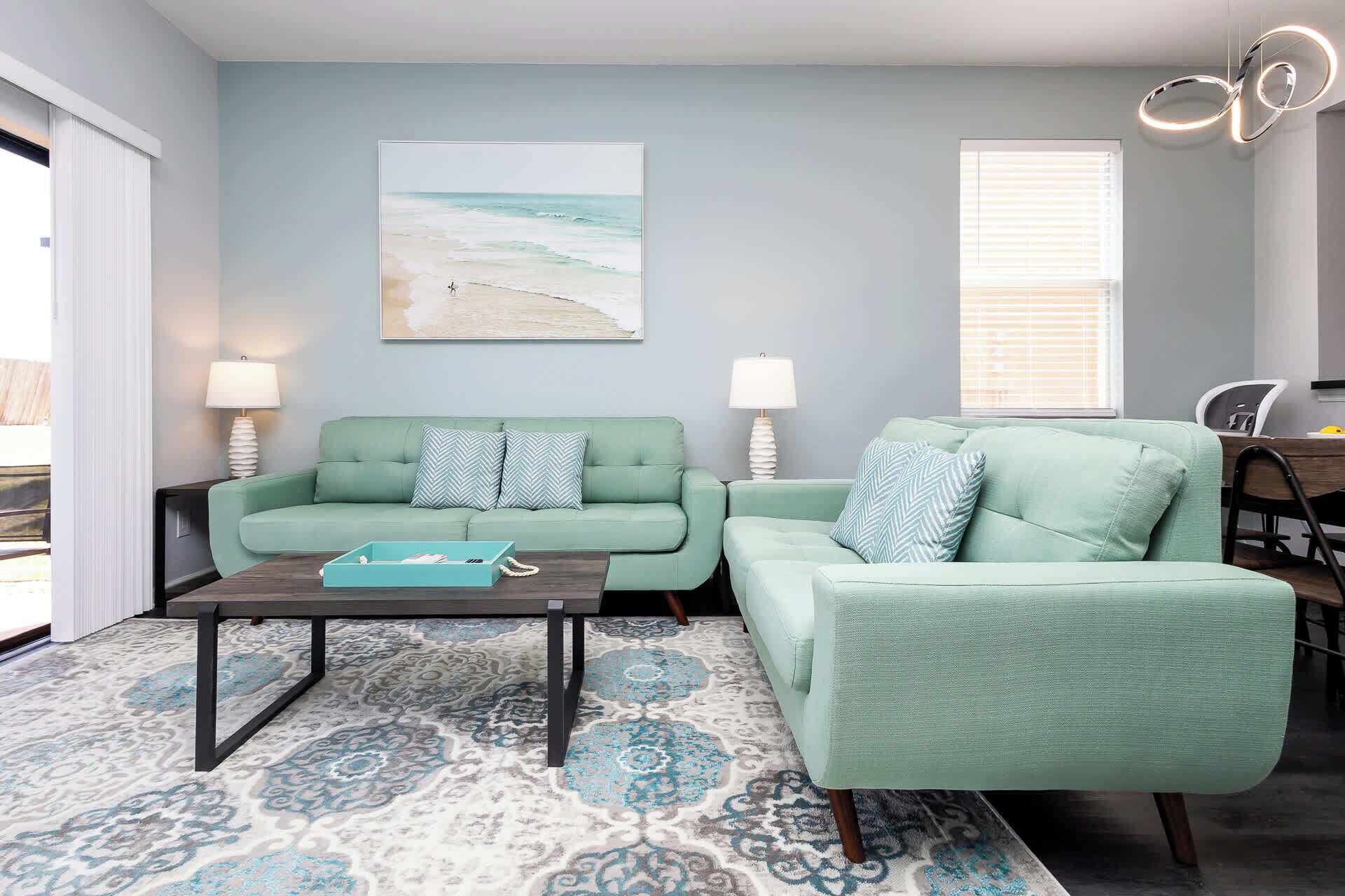 A living room with two light green sofas, a coffee table, patterned rug, and a beach-themed painting on the wall.