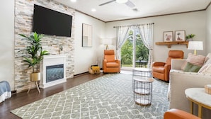 A living room with a fireplace, a flat-screen TV mounted on a stone wall, two leather armchairs, a sofa, a coffee table, and a ceiling fan. - Stone Creek Retreat 2 BR Home! (Branson)