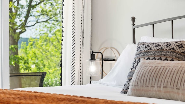 A neatly made bed with patterned pillows, a bedside lamp, and a view of greenery through a window. - Stone Creek Retreat 2 BR Home! (Branson)