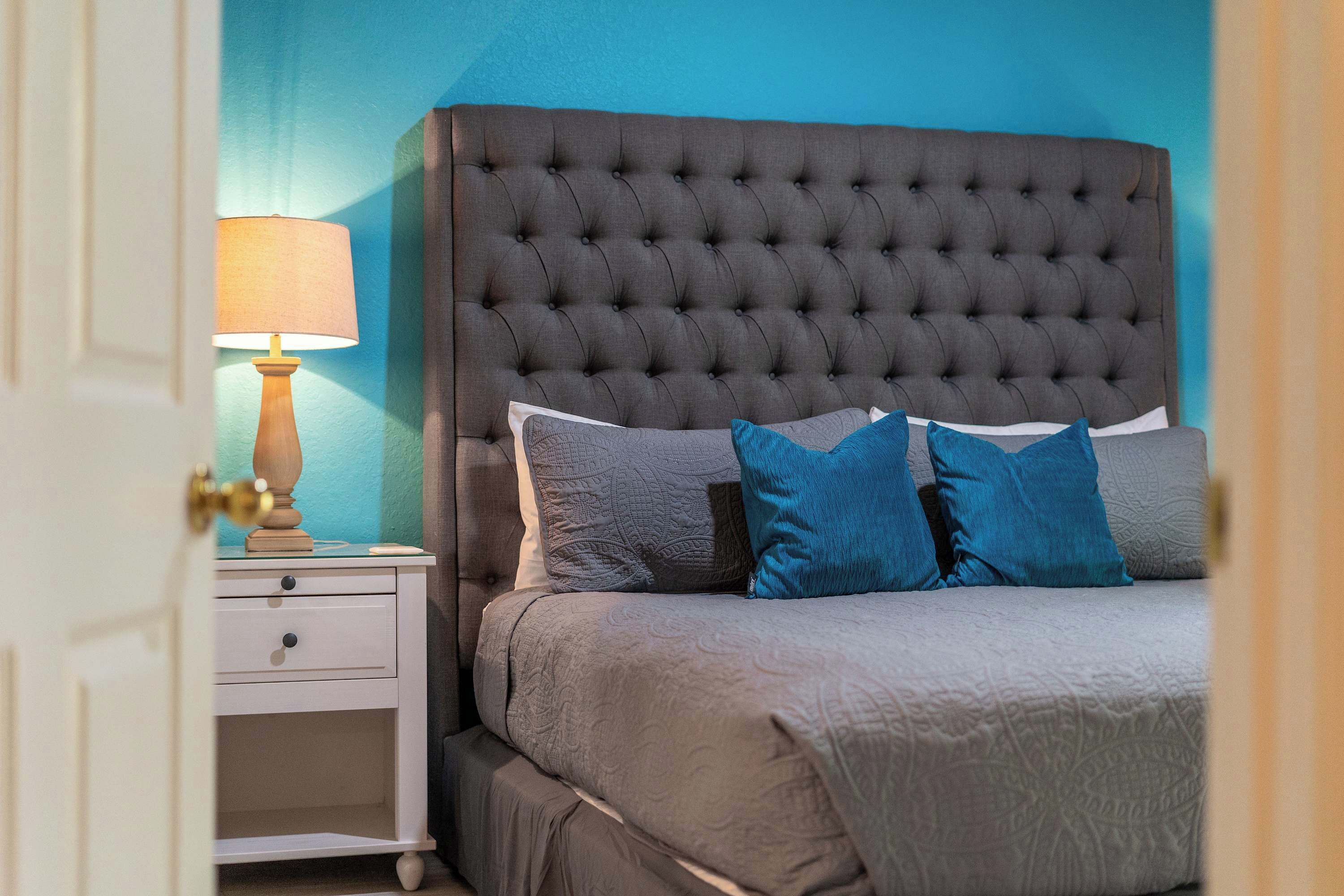 A bedroom with a tufted headboard, a bedside table with a lamp, and a bed with grey bedding and blue pillows.