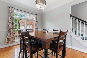A dining room with a large wooden table, six chairs, a chandelier, and a view of a brick house through a window. - Leeloos on the Lake Sleeps 12 (Lexington)