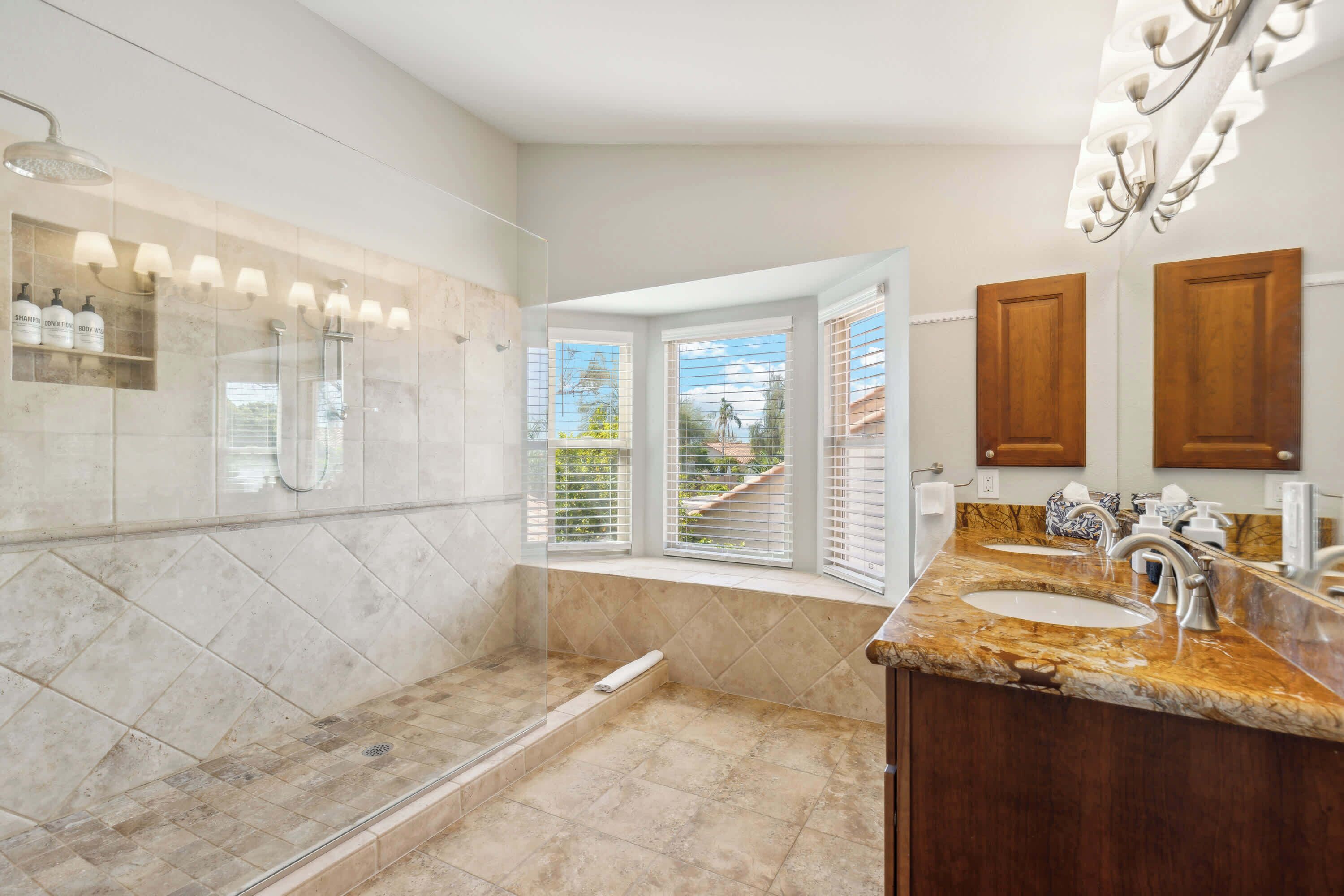 A modern bathroom with a large shower, a vanity with two sinks, and a window with blinds.