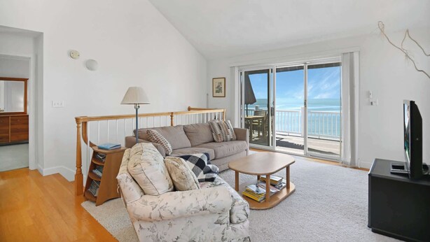 A living room with a sofa, armchair, and coffee table, a TV, and a balcony with a view of the sea. - 3BR Oceanfront on Nubble Cliffs! (York Beach)