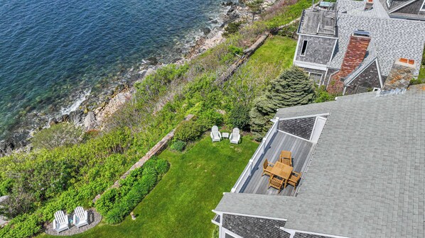 An aerial view of a coastal property with a grassy lawn, two Adirondack chairs, and a wooden deck.