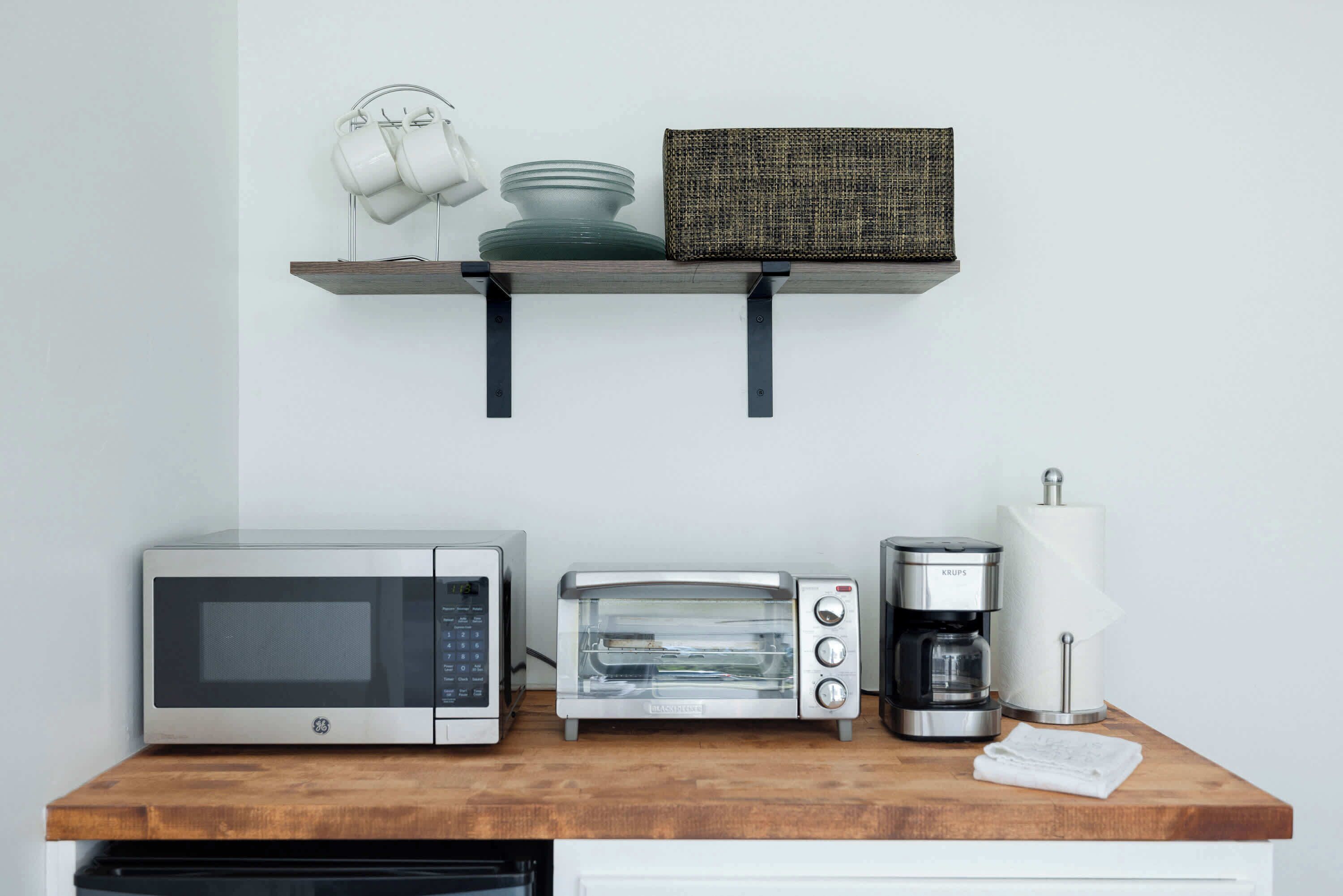 A kitchen with a microwave, toaster, coffee maker, and a shelf with cups and a basket.