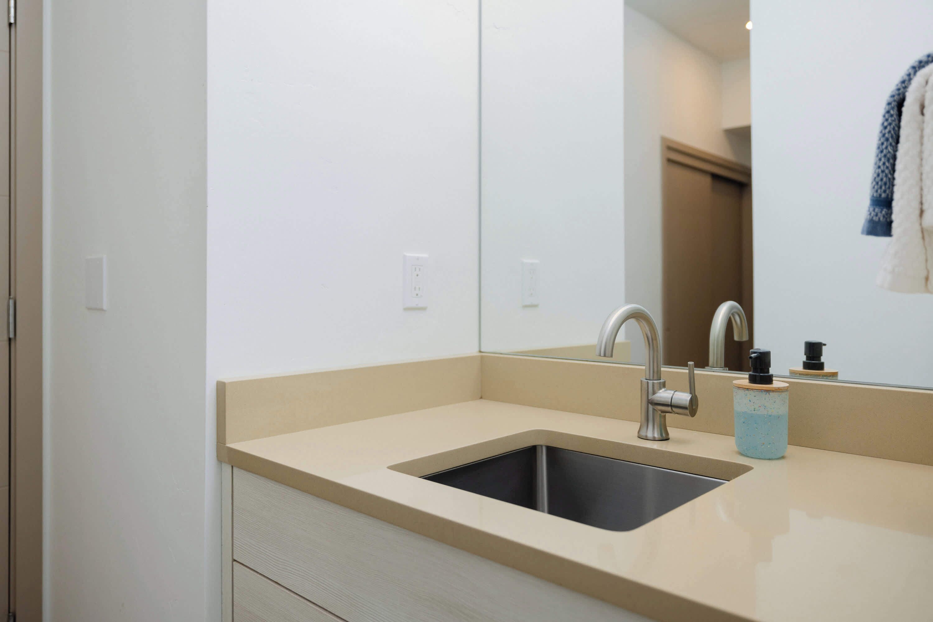 A modern bathroom with a beige countertop, a stainless steel sink, and a mirror reflecting a towel rack and a door.