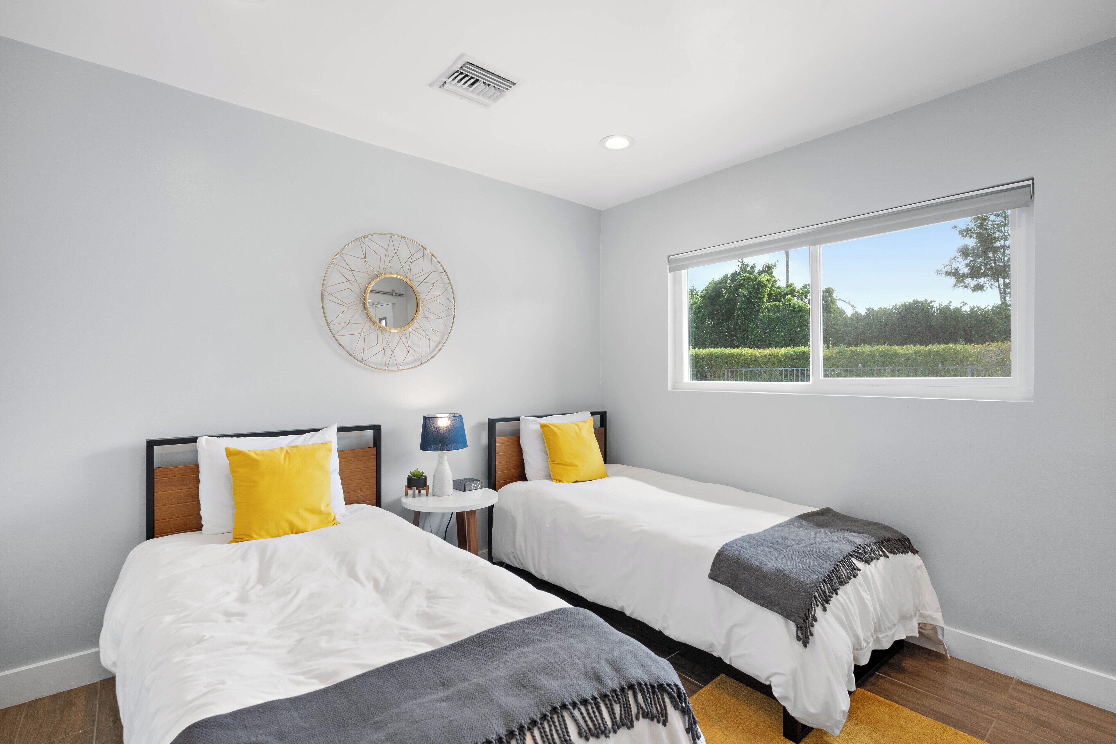 A bedroom with two beds, a round mirror, a window, and a small table with a lamp.