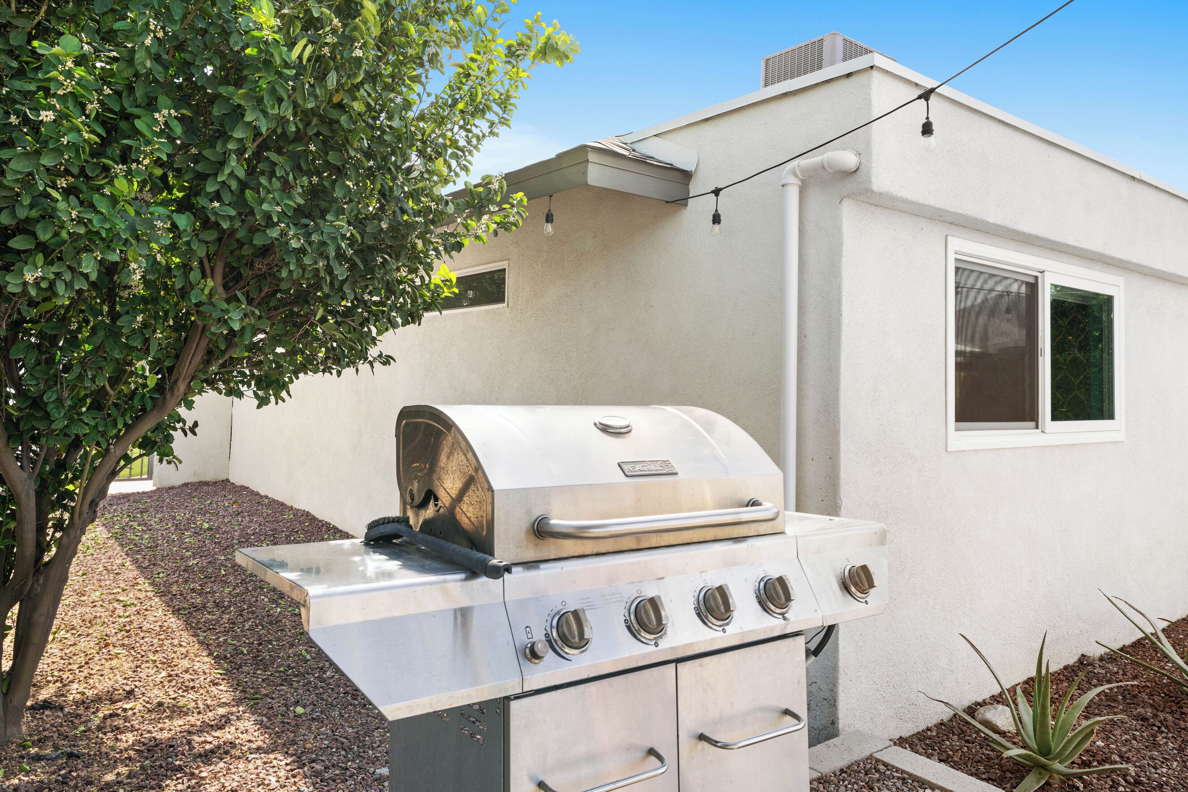 Un barbecue en acier inoxydable doté de quatre brûleurs, d’une cheminée et d’un four intégré, placé devant un bâtiment blanc orné d’une fenêtre et d’un petit arbre sur le côté gauche.