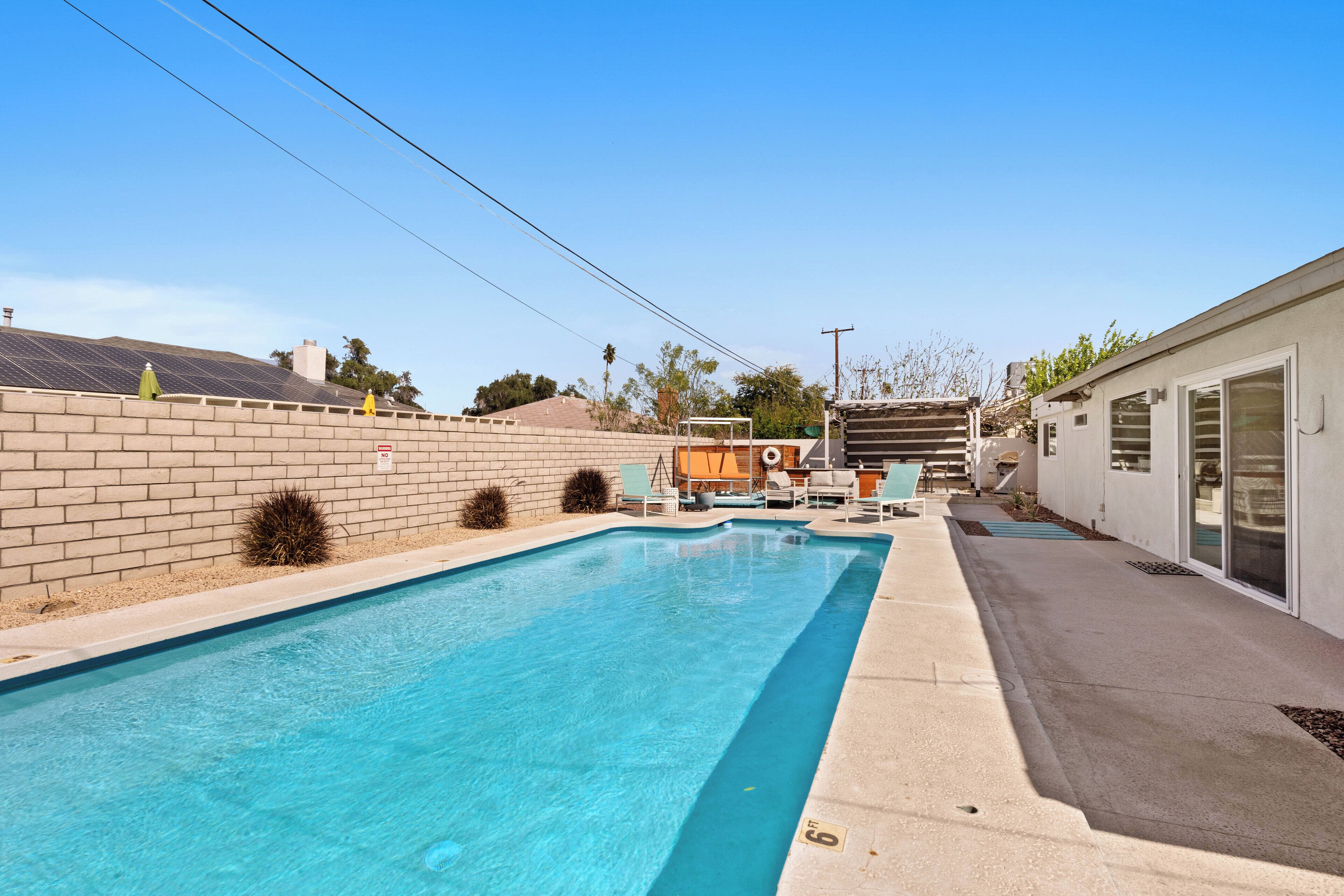 A rectangular swimming pool with a tiled deck, a brick wall, and a covered patio area with outdoor furniture.