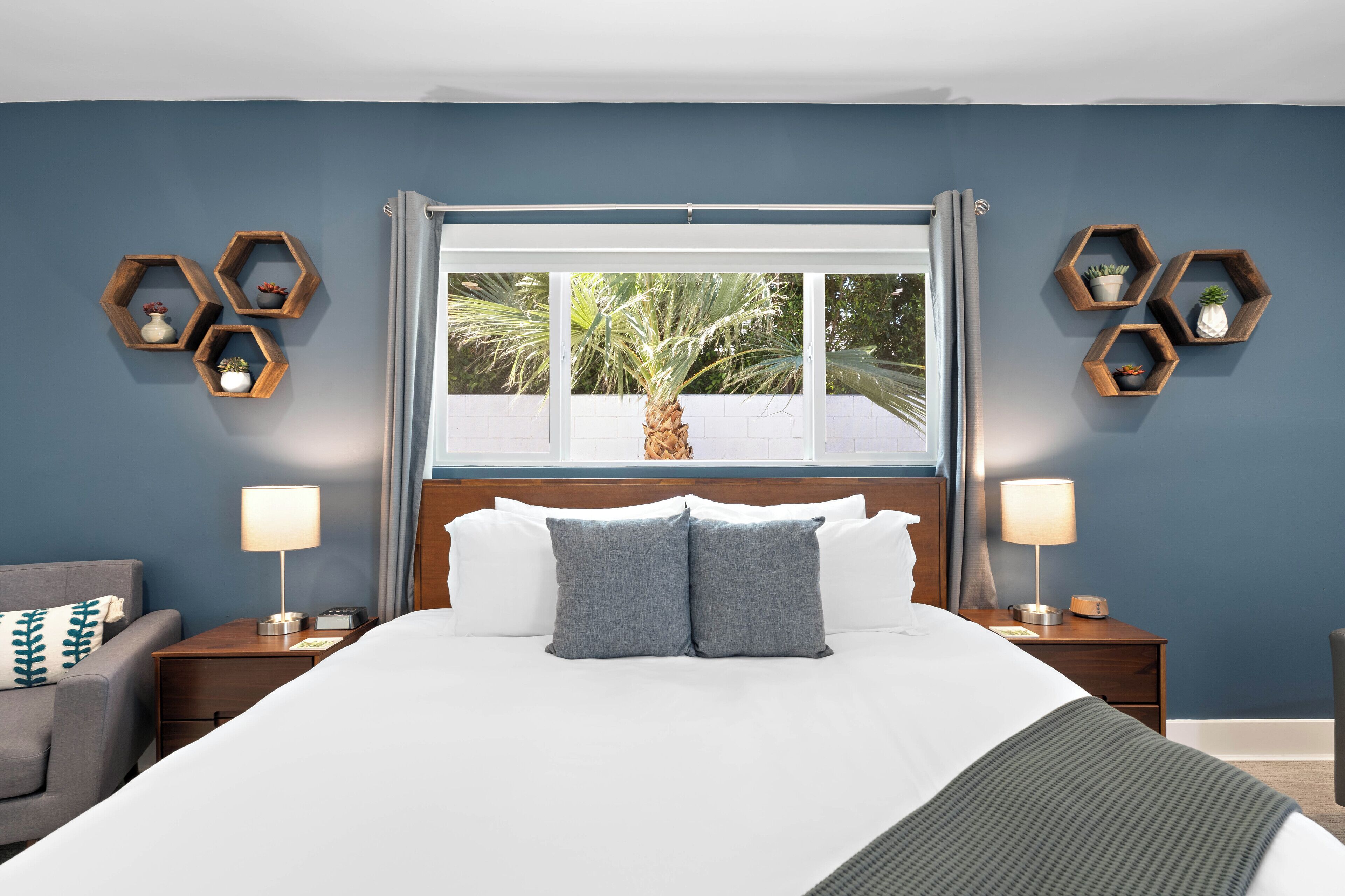 Une chambre d’hôtel avec un grand lit, deux tables de chevet avec des lampes, une table d’appoint, une chaise et une fenêtre donnant sur des palmiers.